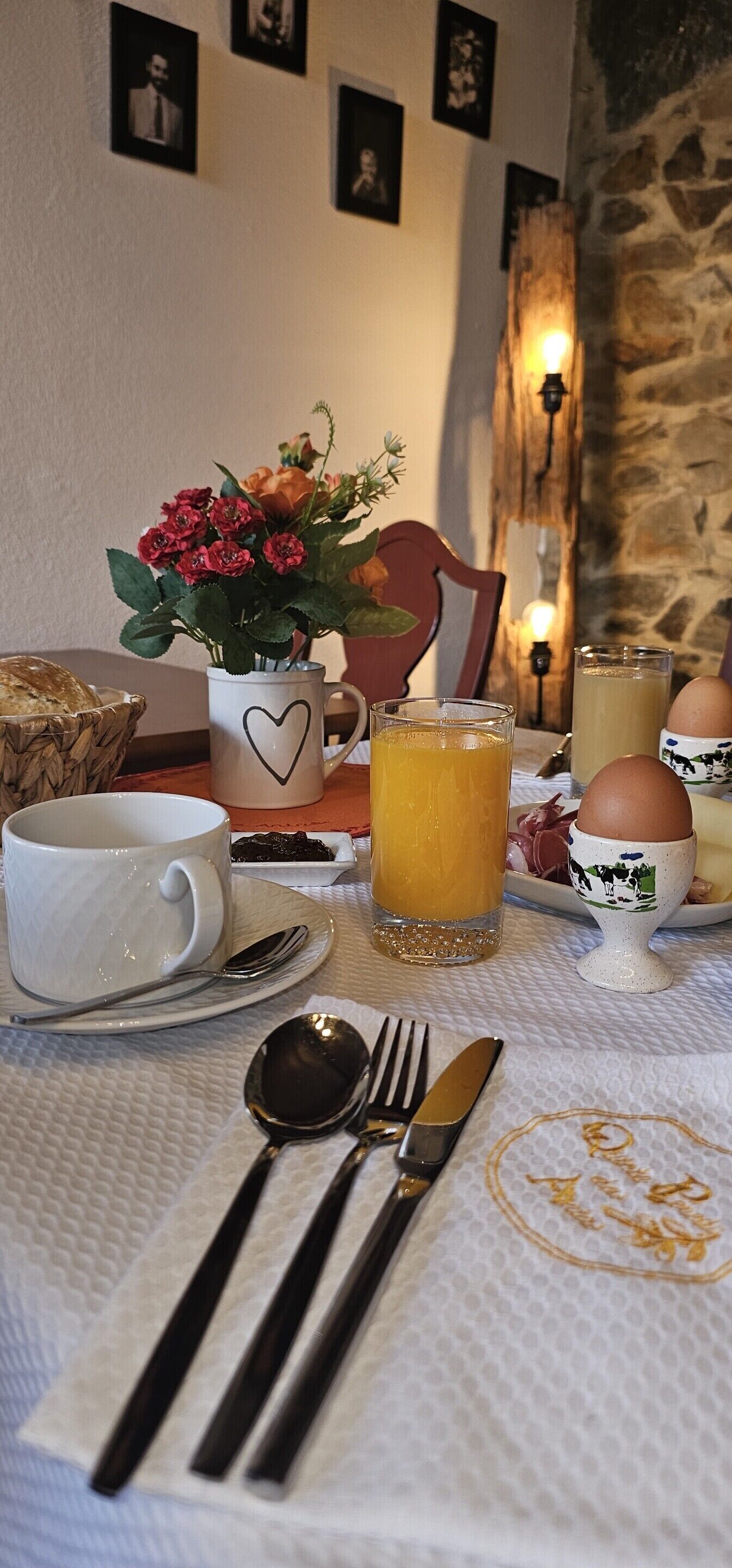 Una escena de desayuno con una mesa preparada para dos personas, que incluye una taza blanca, un vaso de zumo de naranja, un plato con huevos y una cesta de pan.