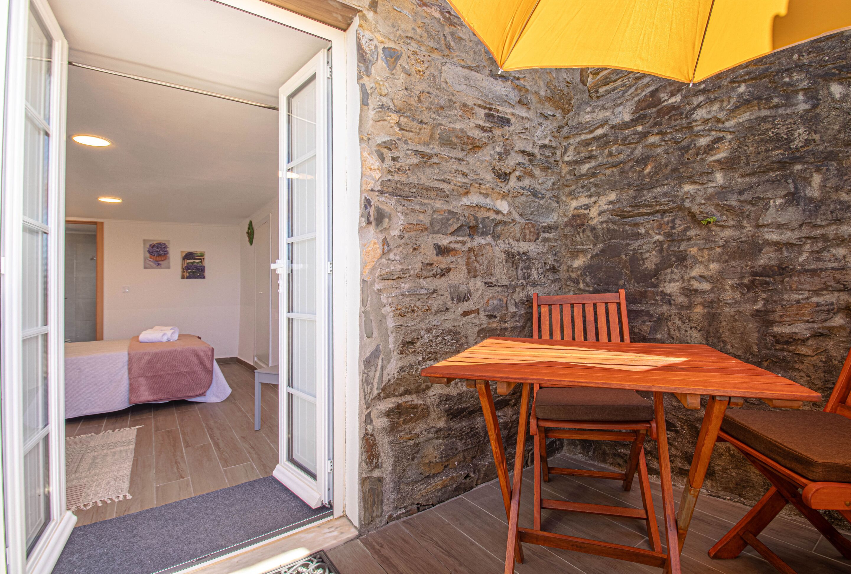 Una habitación con pared de piedra, una mesa de madera con dos sillas, una cama con manta rosa y una puerta que da a otra habitación.