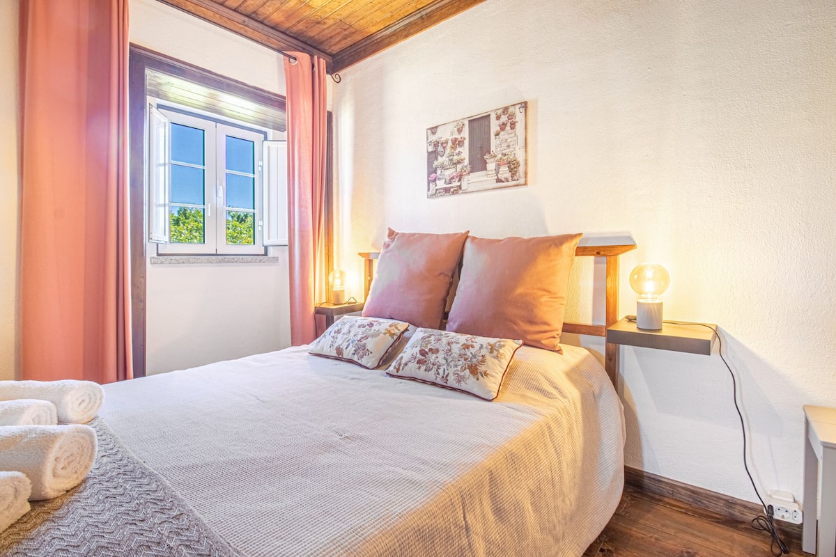 A neatly made bed with floral pillows, a bedside table with a lamp, and a window with curtains.