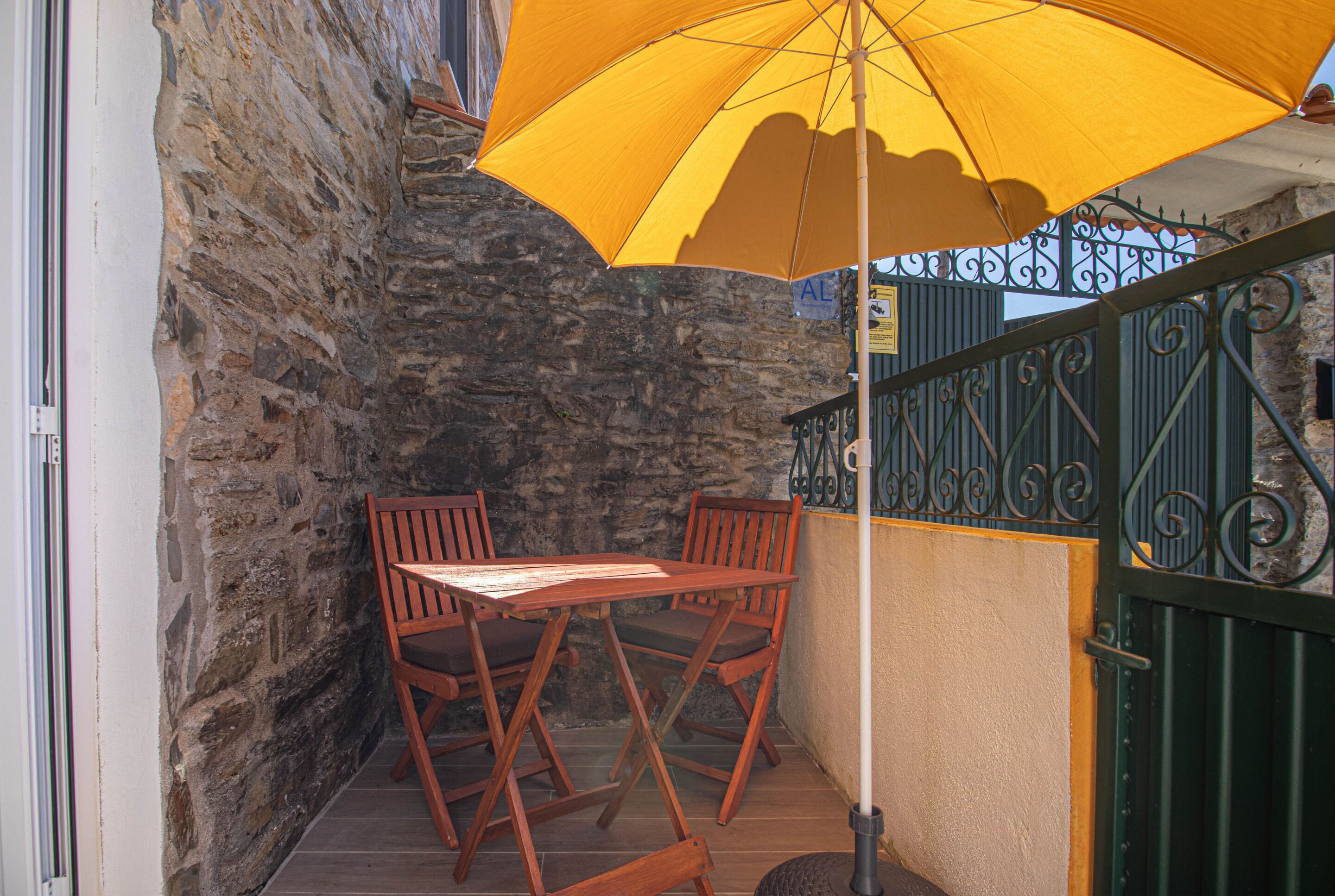 A stone wall, wooden table and chairs, and a yellow umbrella.