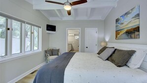 A bedroom with a large bed, a ceiling fan, a painting on the wall, and a view of the outdoors through the windows.