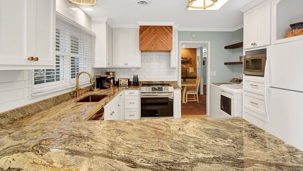 Een moderne keuken met witte kasten, granieten aanrechtbladen en een centraal eiland. Er is een houten backsplash in visgraatmotief, een inbouwoven en een magnetron.