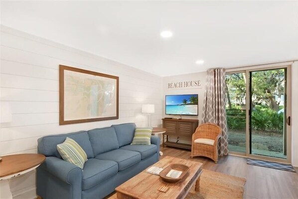 A living room with a blue sofa, a wooden coffee table, a television, and a wicker chair. - Villa By The Sea #254 - Getty Building (Jekyll Island)