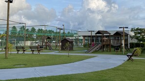 Un parque infantil con toboganes y columpios, rodeado de bancos y un edificio con grandes ventanales.