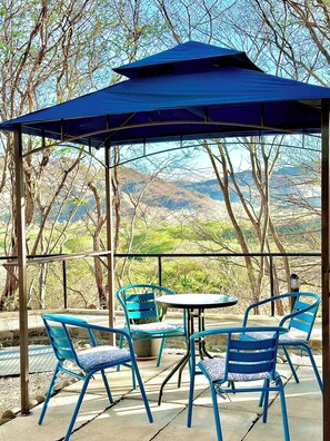 A covered outdoor seating area with blue chairs and a table, overlooking a mountainous landscape. - Ocotal Hilltop Condo: Mountain Views, 4 Pools, 3 Min to Beach, 100 Mbps WiFi (Ocotal)