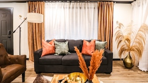 Een woonkamer met een donkere bank, meerdere kussens, een houten salontafel met decoratieve objecten, een vloerlamp en een groot raam met gordijnen.