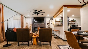 Een moderne woonkamer met een open haard, een eethoek met een houten tafel en stoelen, en een keuken met planken en een magnetron.
