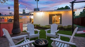 A backyard with a fire pit, white Adirondack chairs, and a small building with string lights.