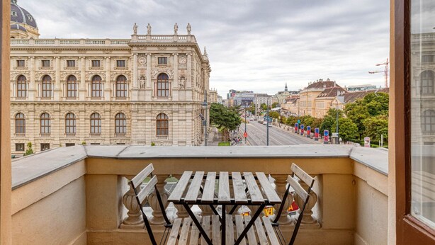 En balkon med udsigt over en historisk bygning med prangant arkitektur, en gade med biler og en klar himmel.