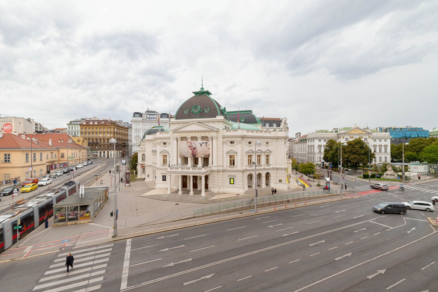 A large, historic building with a prominent dome and classical architecture, located at a busy urban intersection with tram lines and pedestrian crossings.