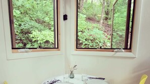 Un comedor con una mesa redonda, dos sillas amarillas y dos ventanas con vistas a un bosque.