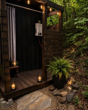 A wooden outdoor shower with a built-in heater, surrounded by rocks and pebbles, with a potted fern and candles for ambiance.