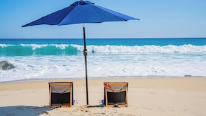 Two beach chairs under a blue umbrella on a sandy beach with waves in the background. - Stunning 11-Bd Villa, Indoor Tennis & Beach Club — Chef, Butler & Yacht Incl. (San Jose del Cabo)