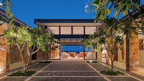 A modern building with a covered entrance, stone walls, and tropical plants. - Stunning 11-Bd Villa, Indoor Tennis & Beach Club — Chef, Butler & Yacht Incl. (San Jose del Cabo)
