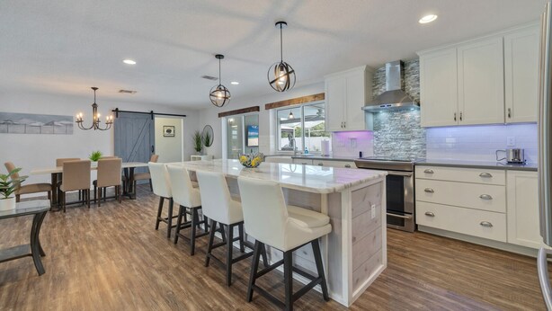 A modern kitchen with white cabinets, a central island, bar stools, pendant lighting, and a stone backsplash.