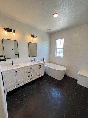 A modern bathroom with a white vanity, two mirrors, a freestanding bathtub, and a window providing natural light.