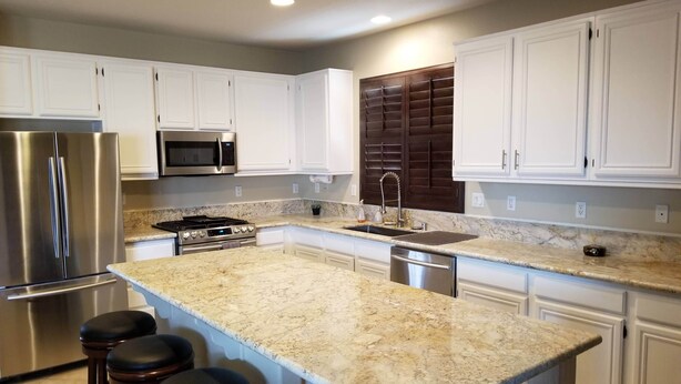 A modern kitchen with white cabinets, a stainless steel refrigerator, a microwave, a gas stove, and a sink. There are three bar stools at the island. - Lancaster Aerospace Basecamp (Lancaster)