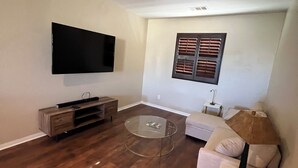 A living room with a flat-screen TV mounted on the wall, a wooden entertainment unit, a glass coffee table, a white sofa, and a window with blinds. - Lancaster Aerospace Basecamp (Lancaster)