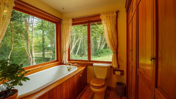 A wooden bathroom with a bathtub, toilet, and large windows offering a view of the forest. - Chalet In Trinidad: There is no place quite like it! (Trinidad)