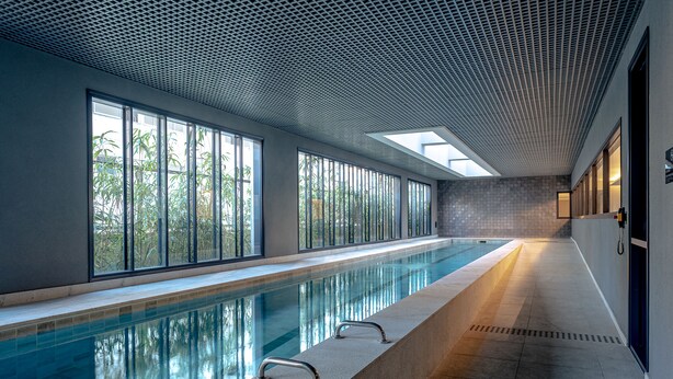 A long indoor swimming pool with clear glass walls, a tiled floor, and a ceiling with a grid pattern. - Studio com piscina aq. na Bela Vista (São Paulo)