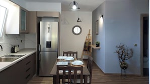 Une cuisine moderne avec un réfrigérateur en acier inoxydable, une table à manger en bois et des chaises. Un petit miroir rond est accroché au mur et une plante en pot est posée sur une table d’appoint.