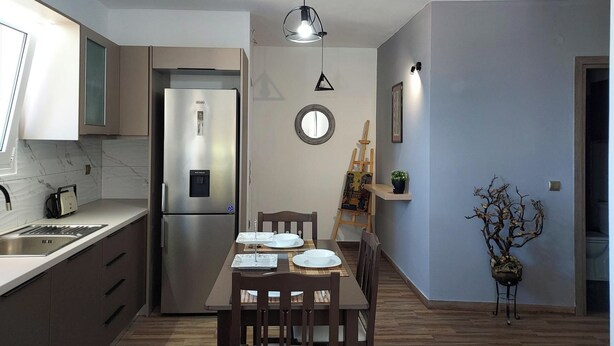 A modern kitchen with a stainless steel refrigerator, wooden dining table, and chairs. There is a small round mirror on the wall and a potted plant on a side table.