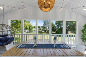 A covered outdoor deck with a wooden table, two chairs, a hanging chair, and a large window overlooking a lake.