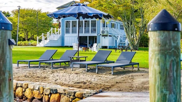 Una casa frente a la playa con un porche cubierto, una escalera y sillas de descanso en una zona arenosa.