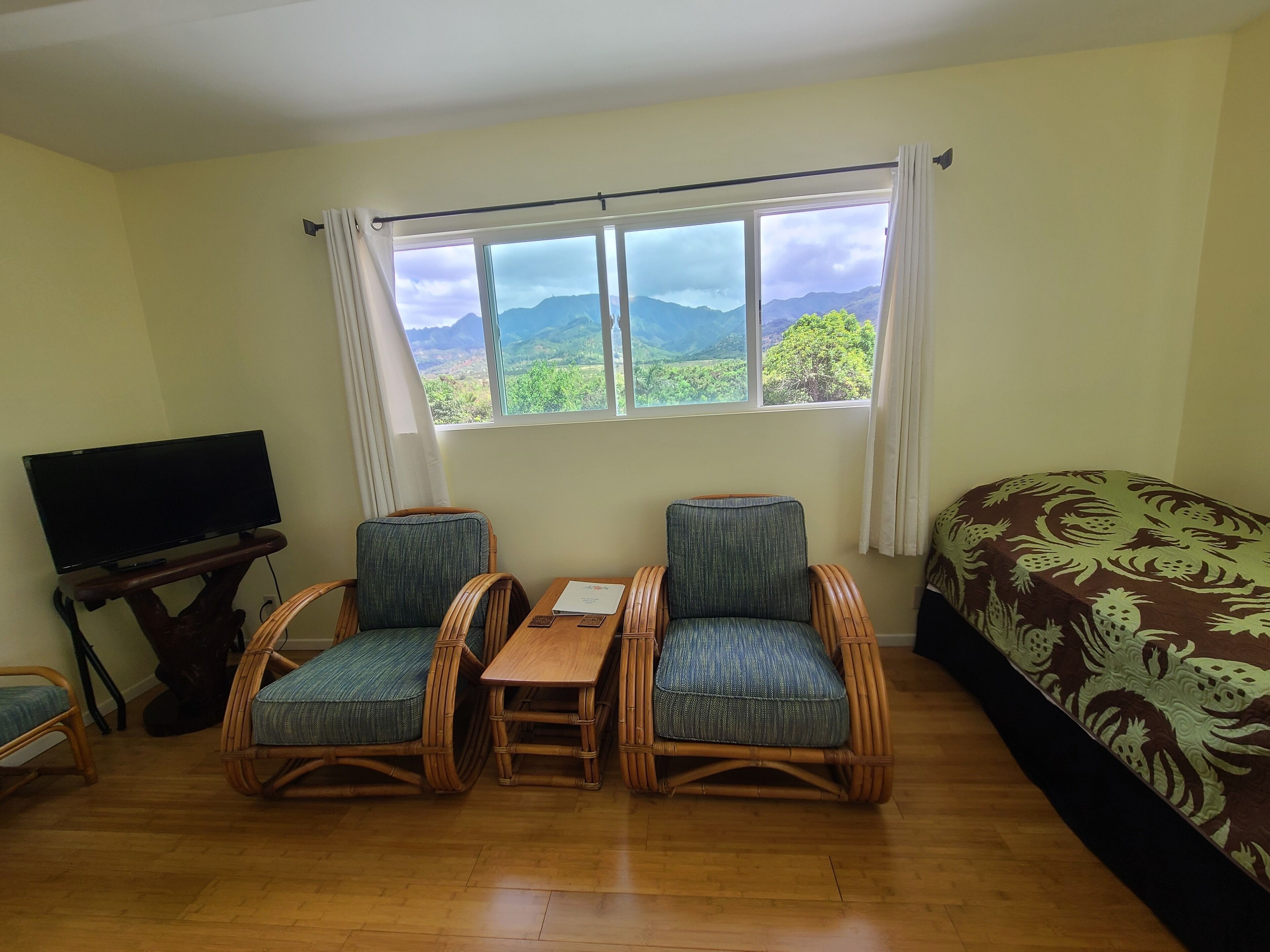 Una stanza con un letto, due poltrone in vimini, un tavolino, una televisione su un mobile e una finestra vista montagna.