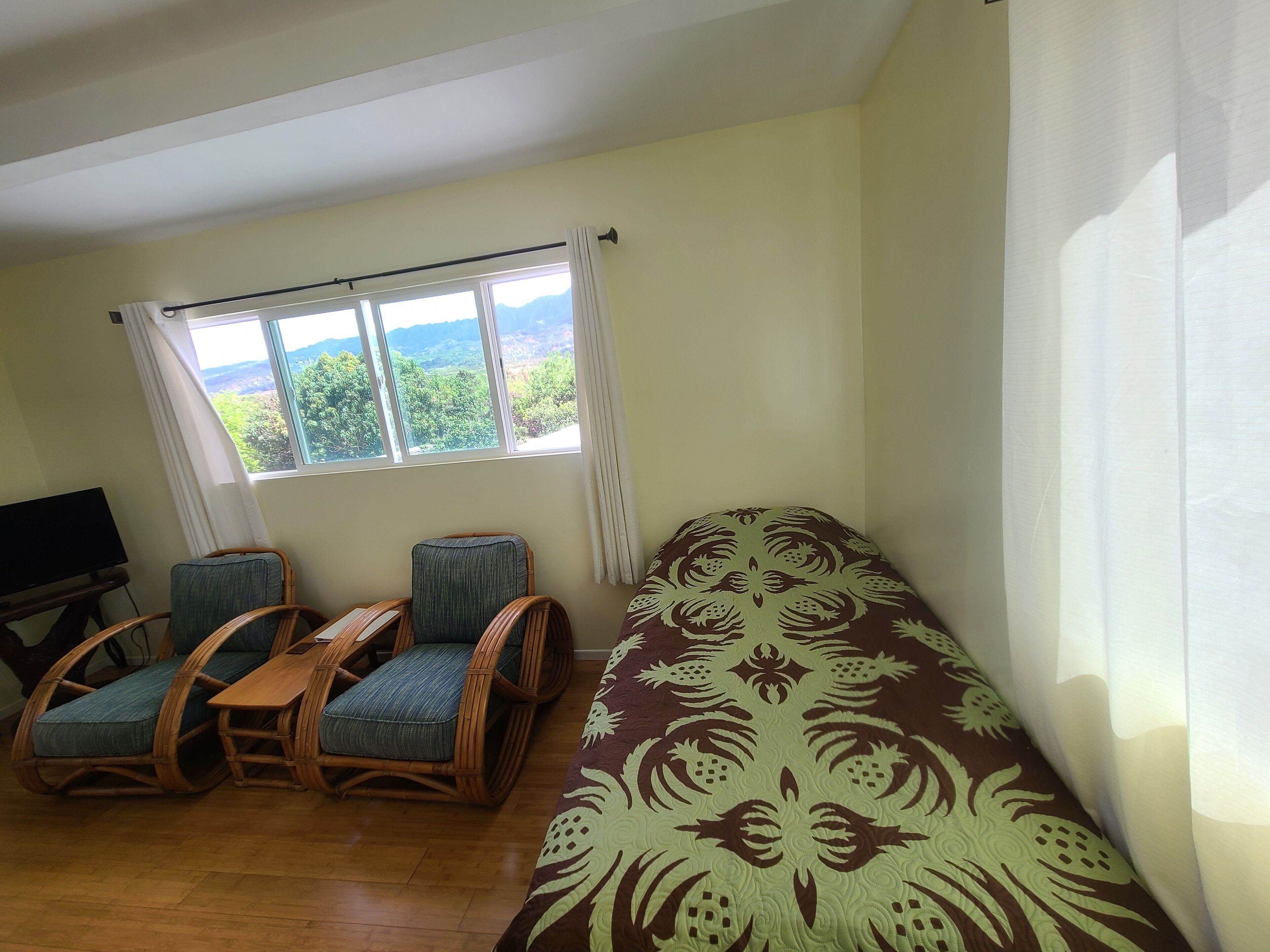 A room with a bed, two wicker chairs, a small table, and a TV.
