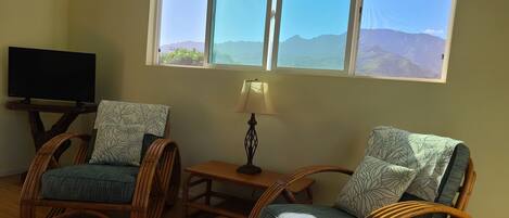 A room with two wicker chairs, a small table with a lamp, and a television on a wooden stand.