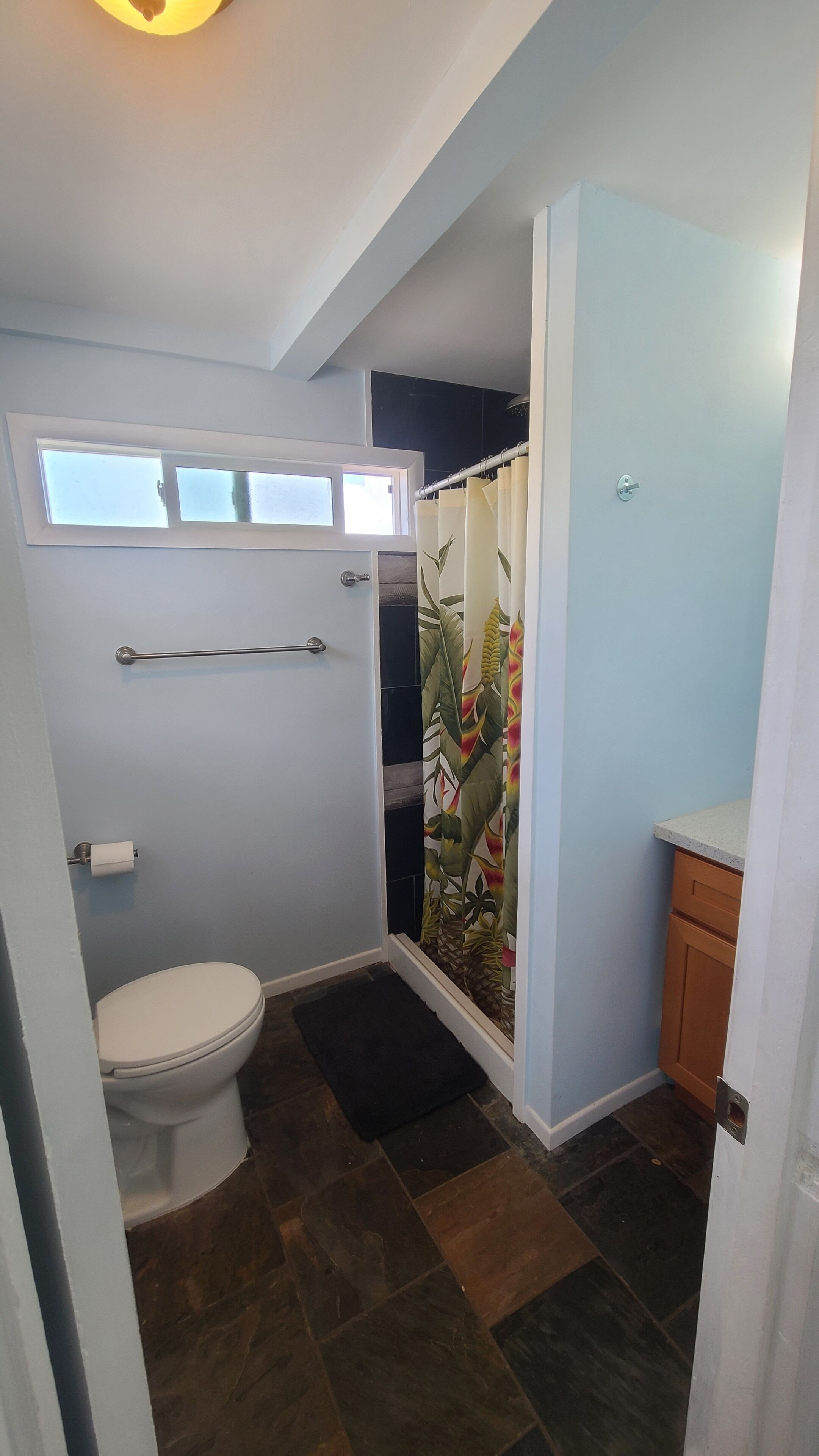 Ein Badezimmer mit Toilette, einer Duschvorhang mit tropischem Blattmuster, einem Handtuchhalter und einem Fenster über der Toilette.