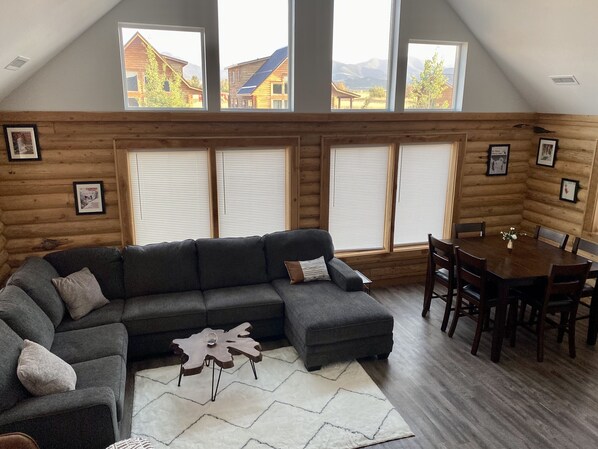 A log cabin interior with a sectional sofa, dining table, and large windows offering views of surrounding buildings and mountains. - Cozy Lazy Dog Cabin (Red Lodge)
