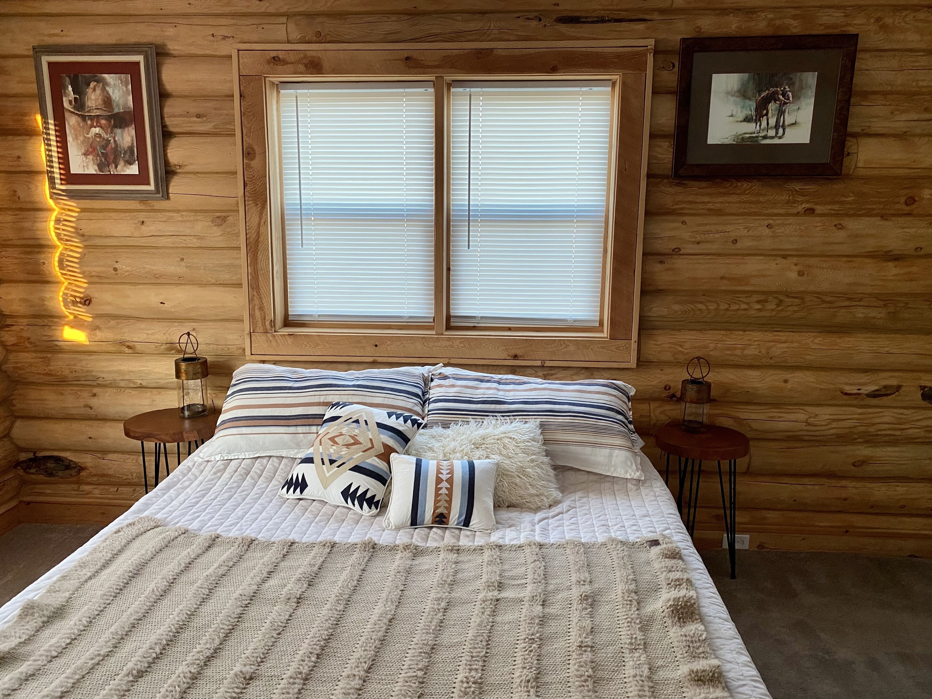 A log cabin bedroom with a large window, a bed with striped bedding, two bedside tables with lanterns, and framed pictures on the walls.