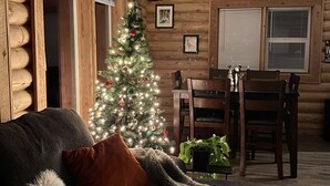 Ein gemütliches Blockhaus-Interieur mit Holzstammwand, Weihnachtstanne, Esstisch mit Stühlen und einer bequemen Couch mit Decke.