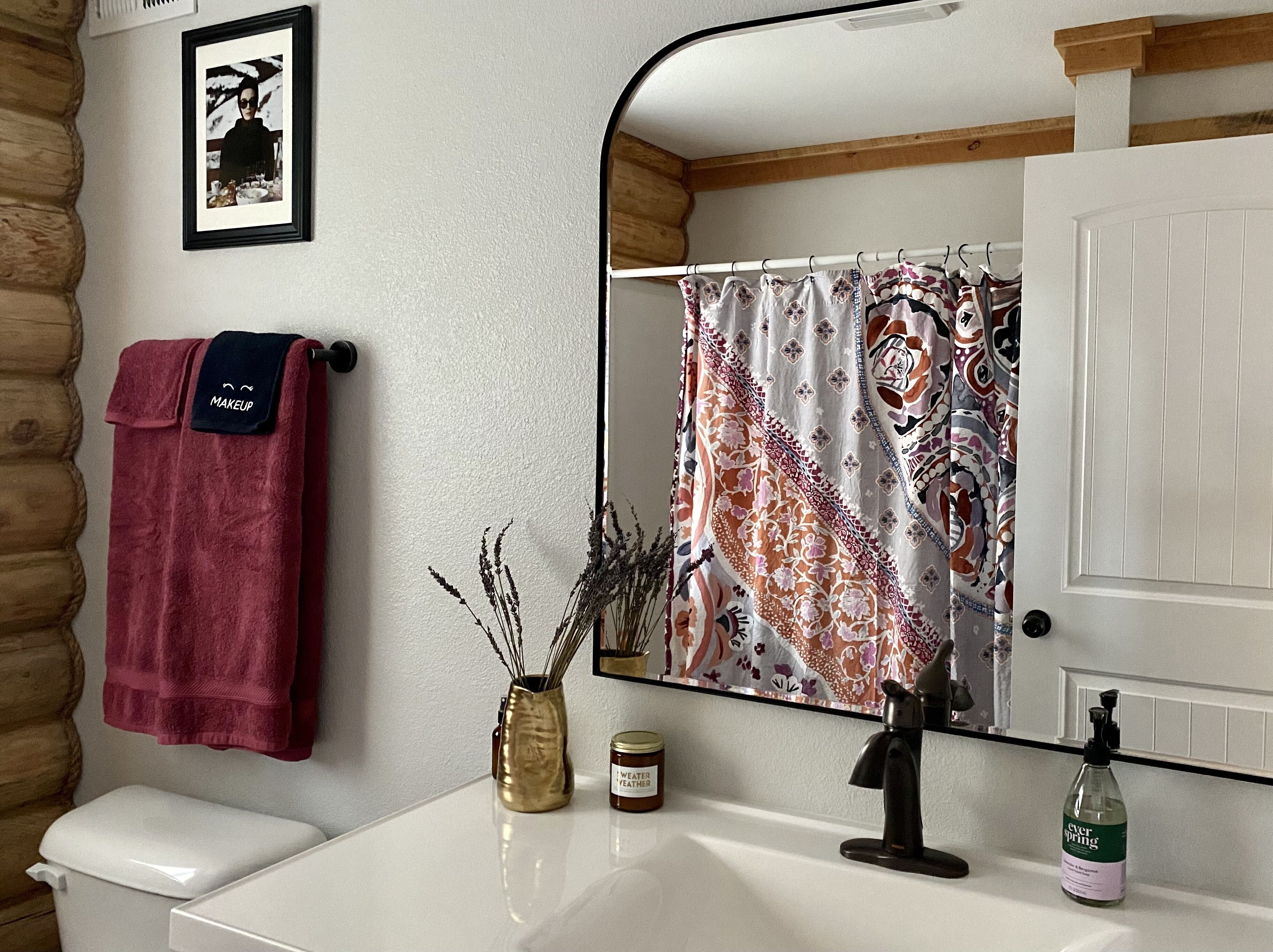A bathroom with a white sink, a mirror, a shower curtain, a towel, a jar of lotion, and a bottle of hand sanitizer.