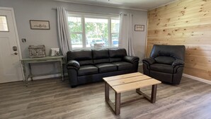 A living room with a wooden floor, a black leather sofa, a black leather armchair, a small wooden table, and a window with curtains.