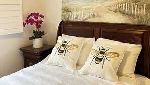 A bedroom with a bed, two pillows with bee designs, a nightstand with a vase of flowers, and a wall painting of a beach scene.