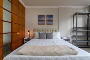 A bedroom with a large bed, a headboard, two bedside tables, a nightstand, a wall-mounted shelf, and a metal shelving unit. - Refuge in Copacabana For 4 People Nsc401 (Rio de Janeiro)