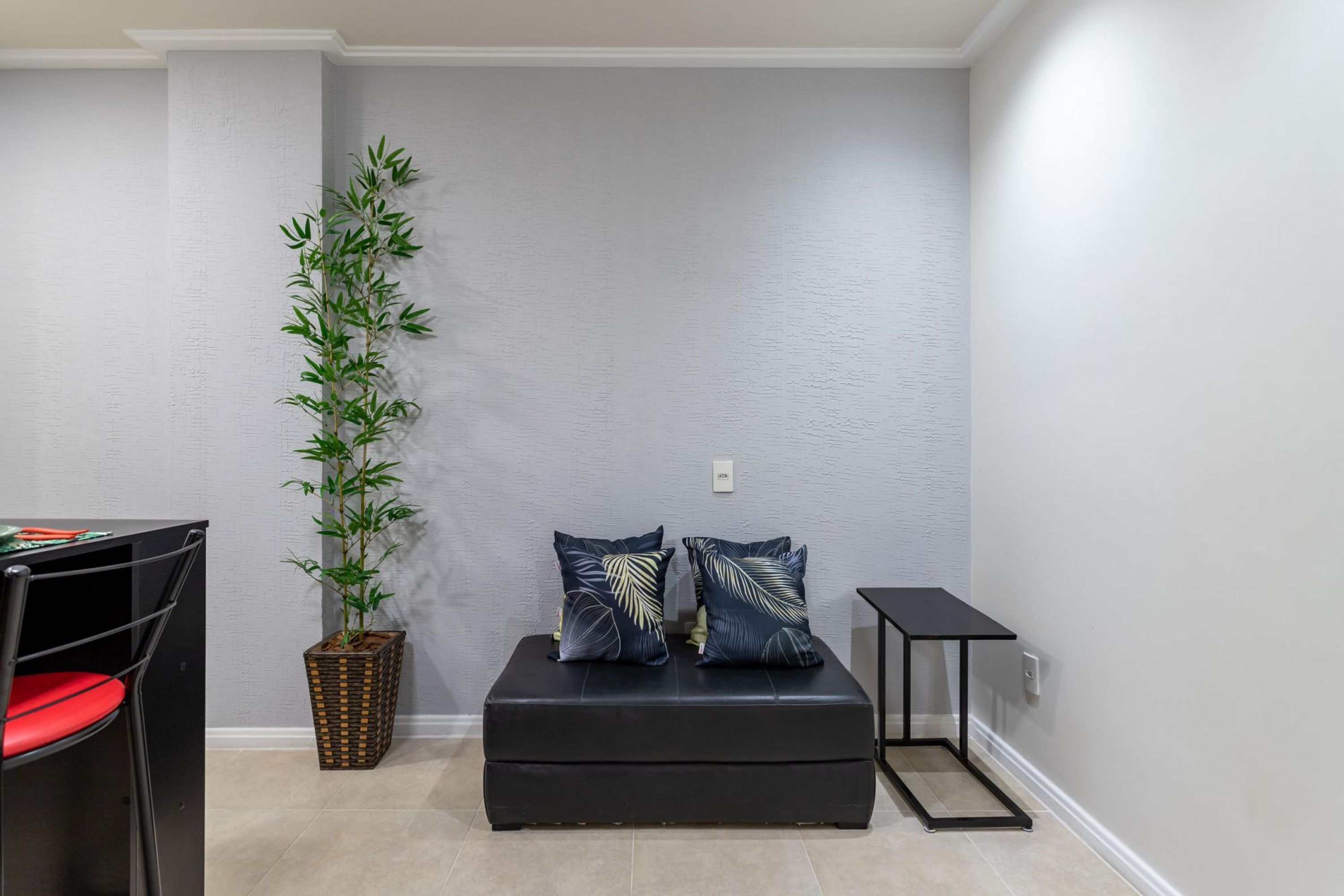 A corner room with a black leather sofa, two decorative pillows, a tall bamboo plant, a small black side table, and a red chair.