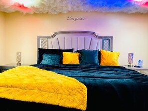 Quarto com cama, almofadas, mesa de cabeceira e parede com desenho de nuvem e texto.