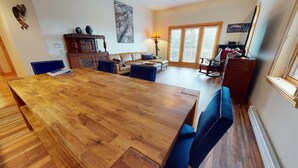 A spacious living room with a large wooden dining table, blue upholstered chairs, a sofa, and a rocking chair. There is a fireplace, a lamp, and a large window with blinds. - Luxury Condo | Close to Lifts | The Grande 117 (Angel Fire)