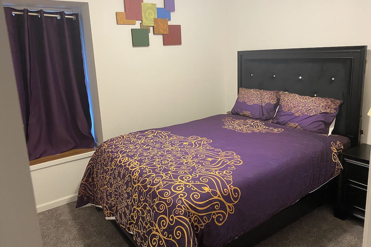 A bedroom with a bed, a headboard, a nightstand, a window with curtains, and a wall decoration.