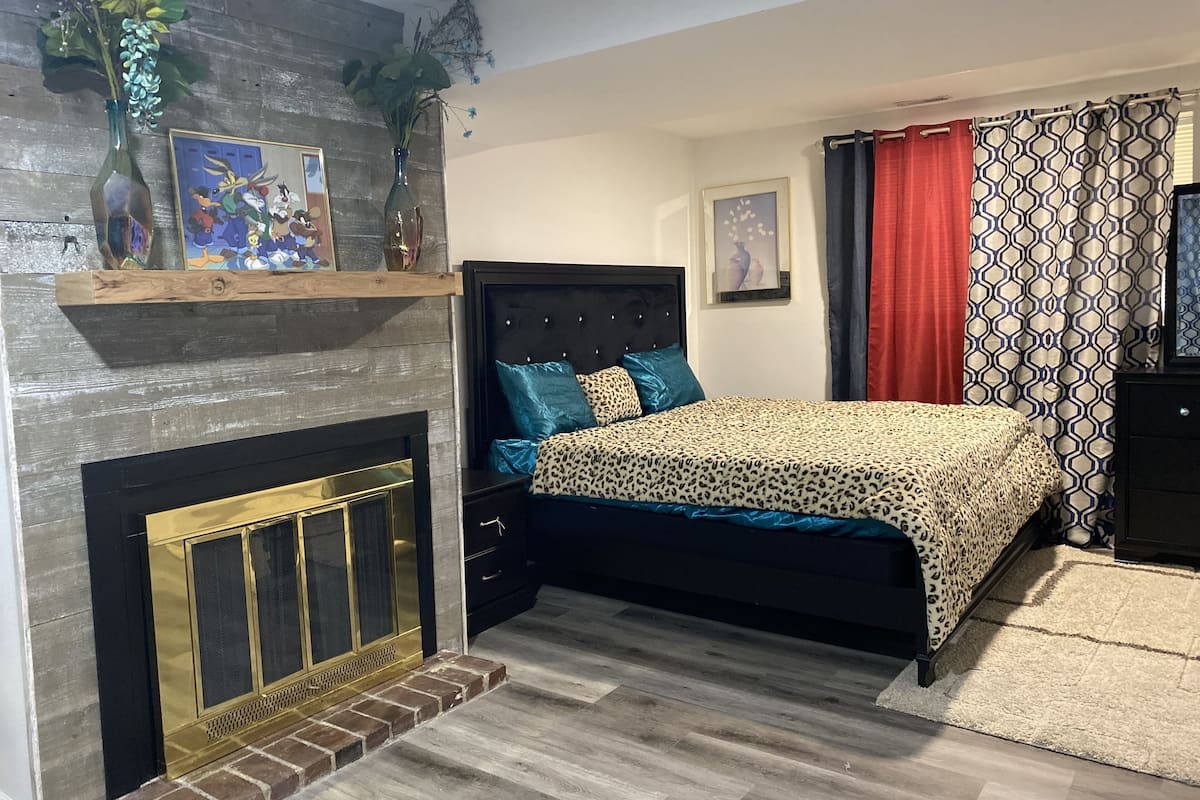 A bedroom with a bed, a fireplace, a shelf with decorative items, and a patterned curtain.