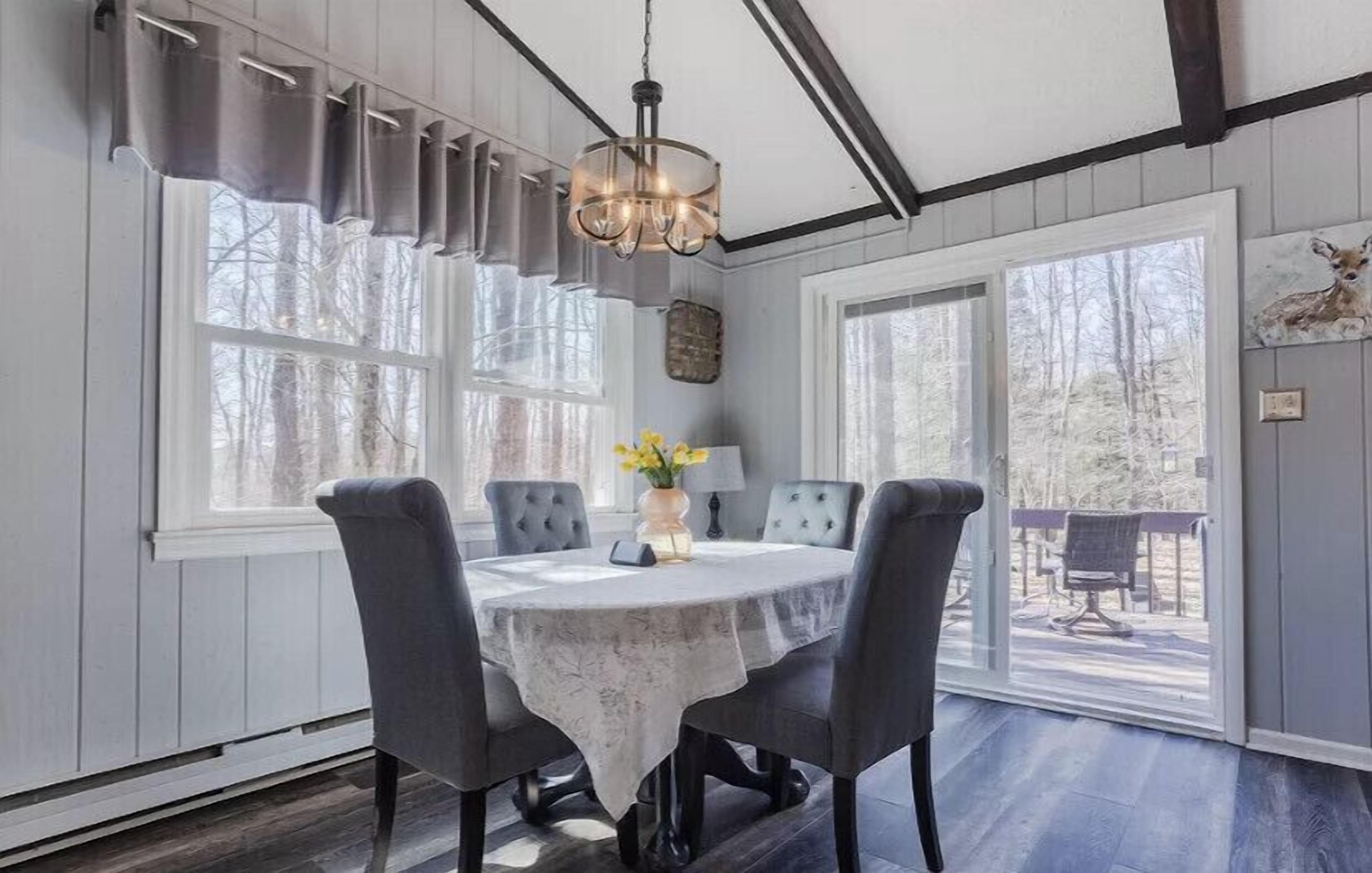 A dining room with a round table, grey upholstered chairs, a chandelier, and large windows with curtains.