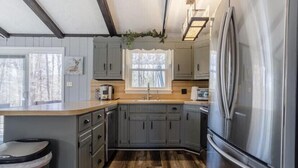 Una cocina con armarios grises, un refrigerador de acero inoxidable, una ventana con vista a árboles y una encimera de madera.