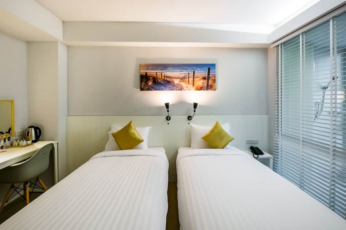 A hotel room with two beds, a desk with a kettle, a phone, and a painting of a beach scene.