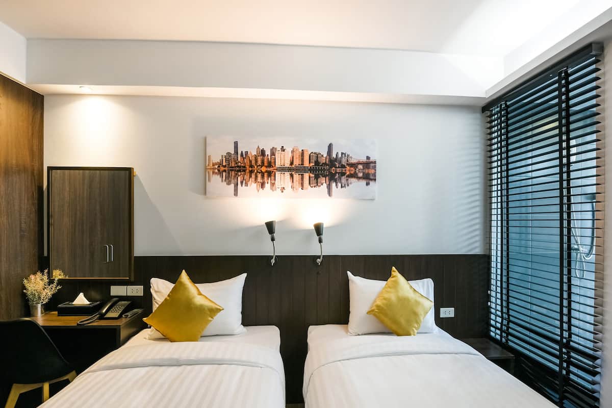 A hotel room with two beds, a cityscape artwork, a desk with a phone, and a window with blinds.