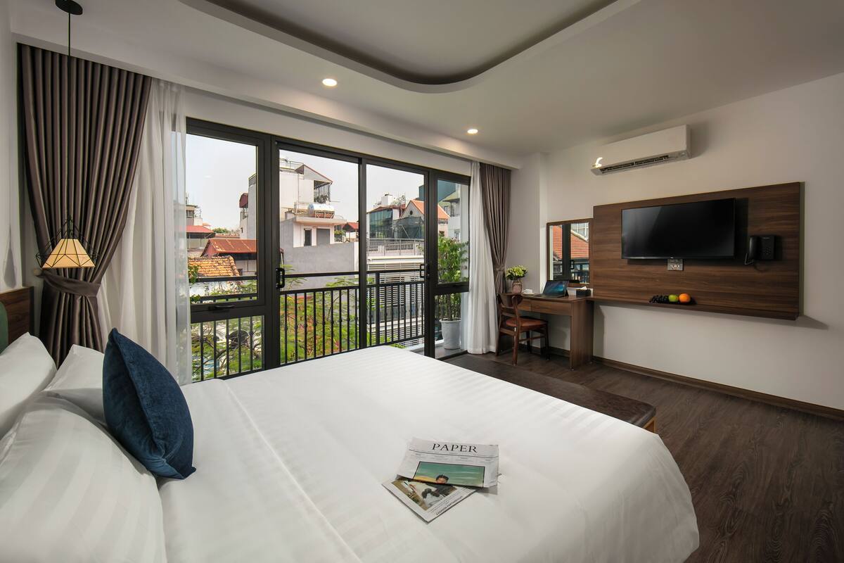 A hotel room with a large bed, a desk, a TV, and a balcony with a view of buildings.