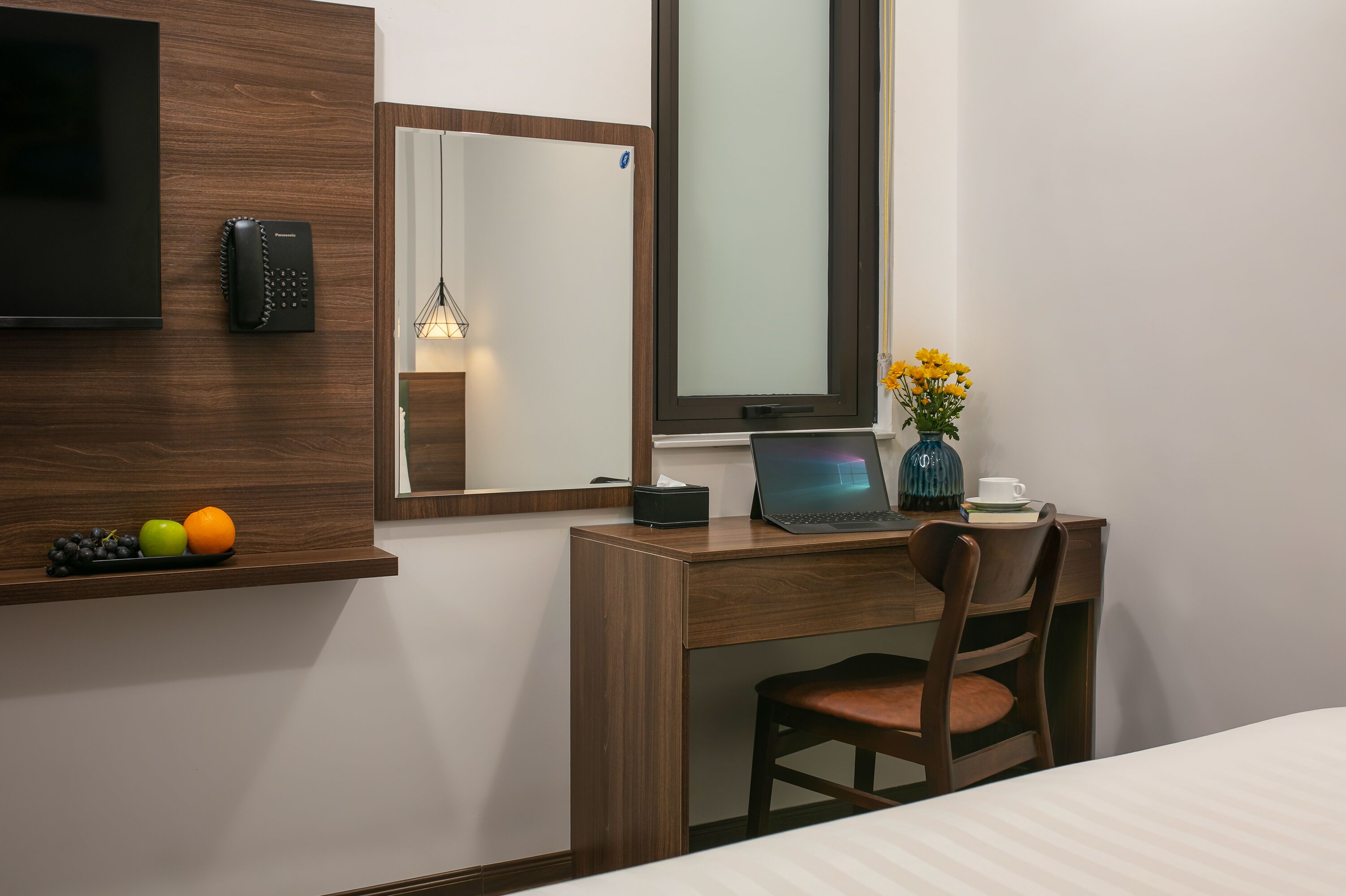 A hotel room with a wooden desk, a laptop, a vase with flowers, and a fruit bowl on the wall.
